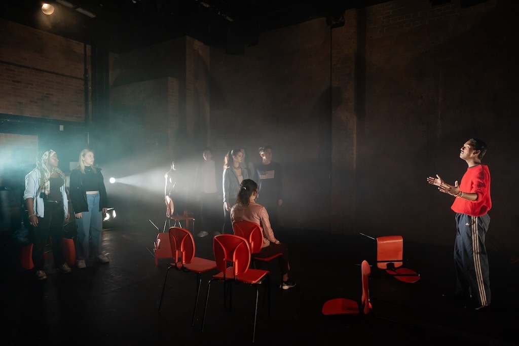 Action shot of the spring 2024 Shakespeare in Performance at RADA final presentation of Julius Caesar. An actor wearing a bright red sweater stands with arms outstretched delivering a monologue, lit by a spotlight. Other students stand or are seated in bright orange chairs.